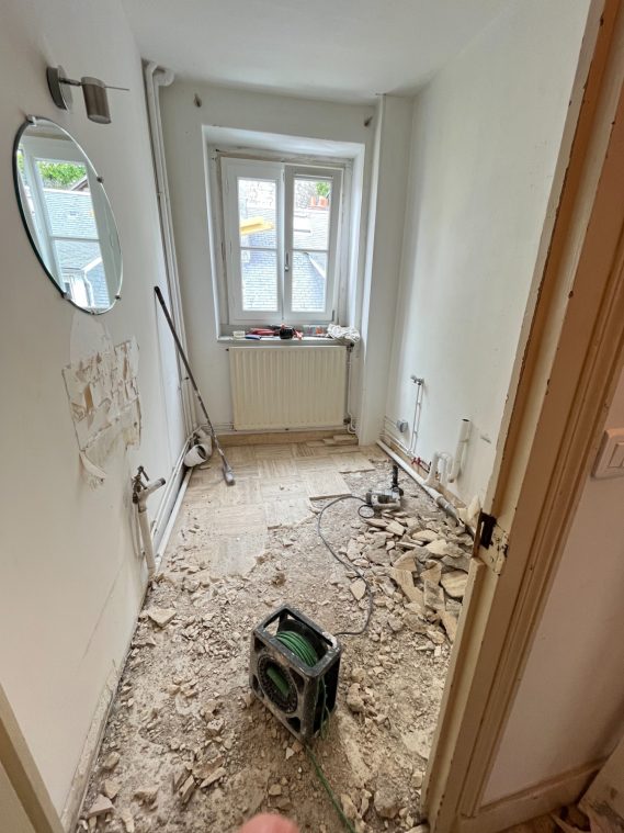 Salle en travaux avec murs nus, sol en désordre et un petit appareil ménager au centre.