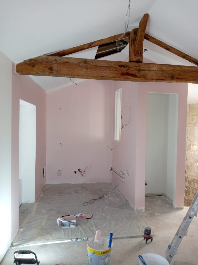 Intérieur d'une pièce avec murs roses, poutres en bois et sol nu. Outils de construction présents.