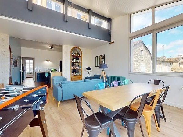 Séjour lumineux avec canapé, table à manger et table de baby-foot. Mur aux fenêtres.