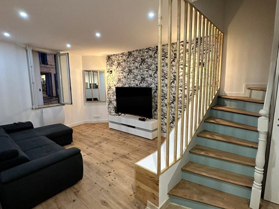 Salon moderne avec un canapé noir, téléviseur, et un escalier en bois menant à l'étage.