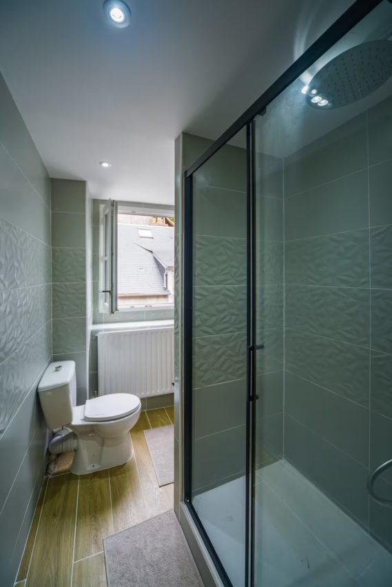 Salle de bain moderne avec douche, toilettes et fenêtre. Mur en carreaux clairs.
