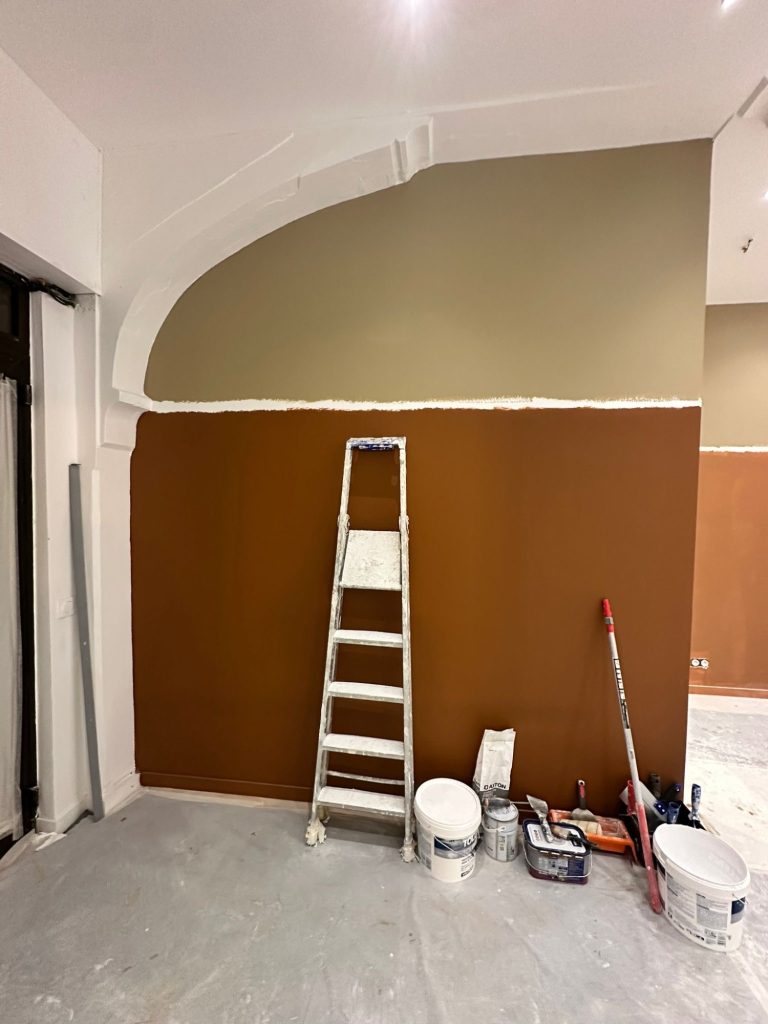 Mur partiellement peint en marron et olive, échelle et matériel de peinture au sol.