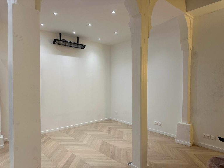 Salle vide avec un sol en parquet, éclairage encastré et murs blancs.